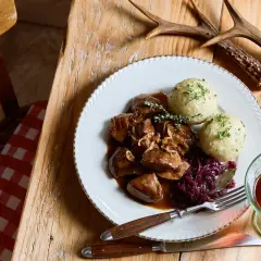 Piatto di cinghiale in dolceforte con canederli e cavolo rosso. Un bicchiere di vino rosso e un palco di cervo completano la scena rustica.