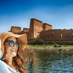 Mujer con sombrero y gafas de sol sonríe en un barco, con templos egipcios de fondo. ¡La mejor época para viajar a Egipto es ahora!
