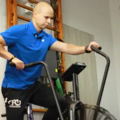 Mężczyzna w niebieskiej koszulce wykonuje ćwiczenia aerobowe na rowerze stacjonarnym, trenując wytrzymałość.