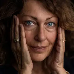 Elisabeth Åsbrink med sina blå ögon och händer mot ansiktet, en bild som väcker tankar om hennes familj och liv.