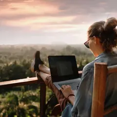 Kobieta z nogami na balustradzie, pracująca zdalnie na laptopie na tle zachodzącego słońca i tropikalnego krajobrazu.