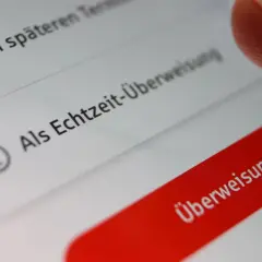 Finger tippt auf "Überweisung prüfen" auf einem Smartphone. Die Option "Als Echtzeit-Überweisung" ist sichtbar. Banken buchen oft sofort.