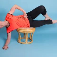 Ältere Frau in orangefarbenem Shirt und schwarzen Leggings macht eine seitliche Übung auf einem Hocker. Ihr linker Unterschenkel ist angewinkelt, der rechte gestreckt.