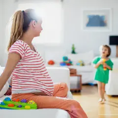 Schwangere Frau sitzt auf Sofa, Kind spielt. Frage: haushaltshilfe nach op wie viele stunden?