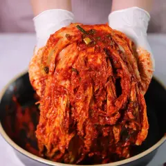 Ręce w białych rękawiczkach trzymają głowę kapusty pokrytą czerwoną pastą. To właśnie kimchi co to jest, tradycyjna koreańska potrawa.