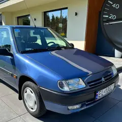 Niebieski Citroen Saxo z niskim przebiegiem, zaparkowany przed nowoczesnym domem. Na desce rozdzielczej widać licznik z przebiegiem 14649 km.