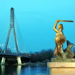 Pomnik Syrenki Warszawskiej z mieczem i tarczą na tle mostu i rzeki.