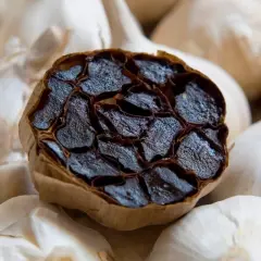 Gousse d'ail noir coupée, révélant ses gousses sombres et moelleuses, entourée d'ail blanc frais. L'ail noir bienfaits et méfaits intriguent.