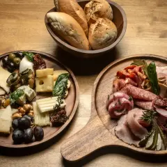 Dwie deski serów i wędlin, pełne smakołyków: sery, oliwki, winogrona, suszone pomidory, wędliny, ogórki i pieczywo.