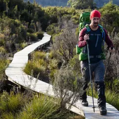 Wanderer mit Rucksack und Stöcken auf Holzsteg. Seine Hose ist mit Gamschen bedeckt, die Schutz vor Gestrüpp bieten.