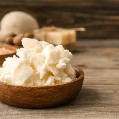 Un bol en bois rempli de beurre de karité, un ingrédient naturel pour les soins de la peau. Des noix de karité et du sel rose sont visibles en arrière-plan.