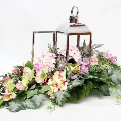 Elegancka kompozycja kwiatowa w lampionie, pełna różowych hortensji, żółtych i zielonych kwiatów, ozdobiona liśćmi i gałązkami.