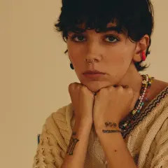 Un'ariete cantante con piercing e tatuaggi, indossa un maglione beige e una collana colorata.