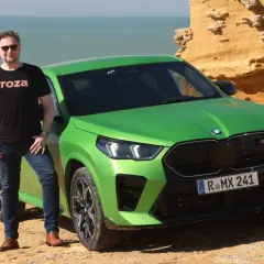 Mężczyzna w czarnej koszulce "Proza" stoi obok jaskrawozielonego BMW X2 M. Samochód zaparkowany jest na skalistym wybrzeżu z widokiem na wzburzone morze.