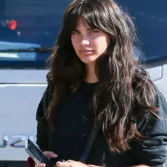 Mujer con cabello largo y oscuro, luciendo un moderno flequillo cortina, mira a la cámara mientras sostiene un teléfono.