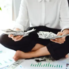 Osoba liczy banknoty dolarowe, przygotowując się do rozliczenia **umowy o dzieło podatek**. Na stole leżą wykresy i pieniądze.