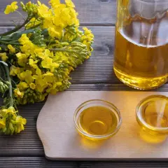Ein Strauß Rapsblüten neben zwei Schälchen und einer Flasche mit goldenem Rapsöl. Rapsöl gesund und vielseitig.