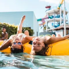 Dwie dziewczyny relaksują się na dmuchanych kołach w basenie. W tle widać zjeżdżalnie wodne, sugerujące, że to największy aquapark w Europie.