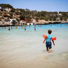 Sonnige Bucht mit vielen Menschen, ein kleiner Junge mit Schwimmflügeln geht ins Meer. Perfekte urlaubsziele mit kindern.