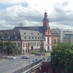 Barocke Kirche mit Turm und Mansarddach, Teil des Jakobsweg Hessen. Autos fahren auf einer Straße, Menschen überqueren sie.