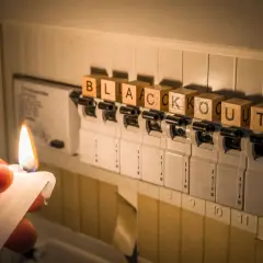 Płonąca świeca oświetla rozdzielnicę elektryczną, na której kostki z literami układają się w słowo "BLACKOUT". Awaria zasilania.