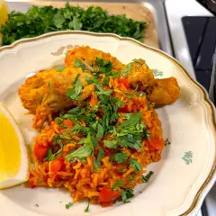 En tallrik med arroz con pollo, kyckling och ris med tomat, garnerad med persilja och citronklyfta. En gryta med samma rätt syns i bakgrunden.