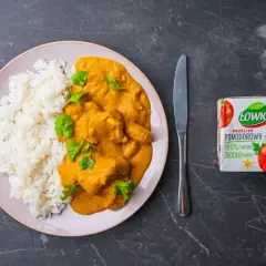 Pyszny butter chicken przepis na talerzu z ryżem, udekorowany świeżą kolendrą. Obok nóż, widelec i przecier pomidorowy Łowicz.