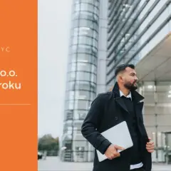 Mężczyzna w płaszczu trzyma laptopa przed nowoczesnym budynkiem.