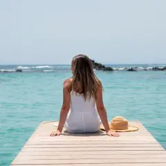 Mujer con vestido blanco sentada en un muelle de madera, disfrutando de la vista al mar turquesa. Su sombrero de paja descansa a su lado, perfecta para viajar sola.
