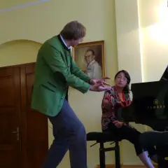 Karolina Nadolska, w zielonej marynarce, gestykuluje do młodej kobiety siedzącej przy fortepianie Yamaha.