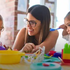 Eine lächelnde Frau mit Brille spielt mit zwei Kleinkindern. Sie lernt den erzieher berufsbegleitend und fördert spielerisch die Entwicklung der Kinder.