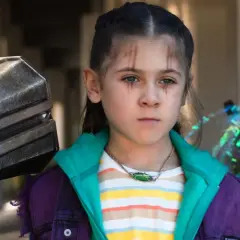 La fille de Thor, avec des marques sur le visage, porte une veste violette et un collier vert. À côté d'elle, le marteau Mjolnir.