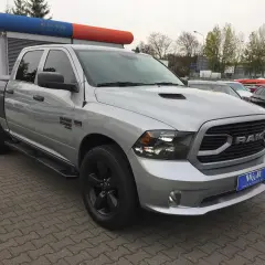 Srebrny Dodge Ram 1500 5.7 Hemi z czarnymi felgami i przyciemnianymi szybami stoi na parkingu przed budynkiem z niebiesko-pomarańczowym banerem.