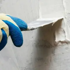 Hand med blå gummihandske spacklar en vägg utomhus. Snickerispackel utomhus används för att jämna till ytan.