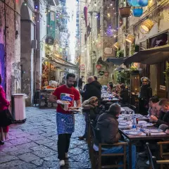 Un vicolo napoletano illuminato da luci colorate, con gente seduta ai tavoli di un locale. Un cameriere porta del cibo.