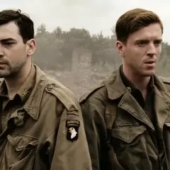 Dwóch żołnierzy w mundurach, symbolizujących bandę braci, stoi na tle zniszczeń. Jeden z nich ma naszywkę "AIRBORNE".