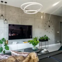 Nowoczesne oświetlenie ściany z telewizorem, z wiszącymi lampami i okrągłym plafonem. Szara ściana z betonową fakturą, czarny telewizor, biała szafka RTV.