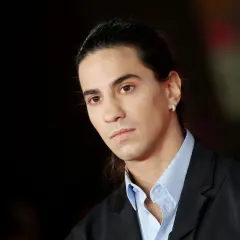 Ritratto di Tedua, rapper italiano, con capelli lunghi legati e un orecchino scintillante. La sua età è difficile da definire, ma emana un'aura di maturità artistica.