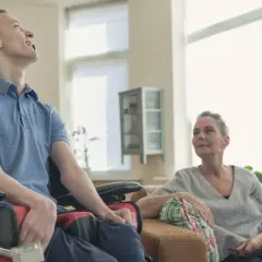 Młody mężczyzna na wózku, zmagający się z chorobą, uśmiecha się do kobiety siedzącej obok.