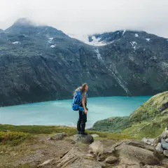 A hiker with a blue backpack stands on a rocky outcrop, overlooking a stunning turquoise lake in Jotunheimen National Park. The dramatic mountains behind are shrouded in mist.