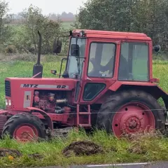 Czerwony ciągnik Belarus 820 pracuje na polu, za nim widać las i zielone pola.