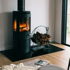 Moderner Holzofen mit brennendem Feuer, daneben ein Korb mit Holzscheiten. Ein gemütlicher Anblick, der an ein Verbot von Holzöfen denken lässt.