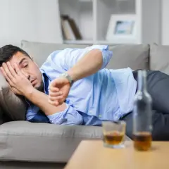 Mężczyzna leży na kanapie, zasłaniając twarz ręką, obok butelka i szklanka alkoholu. Zastanawia się, jak szybko wytrzeźwieć.