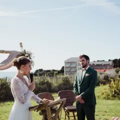 La mariée, en robe blanche, parle au micro lors de la trame cérémonie laïque. Le marié, en costume vert, l'écoute attentivement.