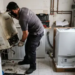 Técnico trabajando para cambiar caldera de gas. Retira una vieja y sucia caldera Junkers, mientras una nueva Vaillant espera en su caja.