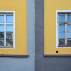 Dwa stare drewniane okna z białymi ramami na żółtej ścianie, rozdzielone szarym pasem.