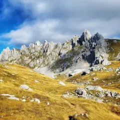 A winding path leads through golden grass and rocks towards the jagged peaks of Bobotov Kuk, a stunning hike under a dramatic sky.