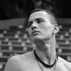 Joven con buzz cut largo, collar plateado y mirada perdida al horizonte.