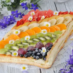 Kolorowa tarta z owocami: kiwi, mandarynki, banany, truskawki, winogrona i borówki, ozdobiona stokrotkami.