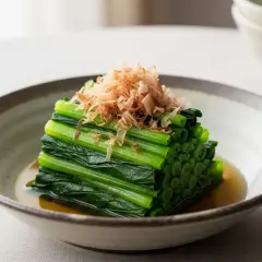 A neat stack of blanched spinach, known as spinach ohitashi, is served in a shallow bowl with a light broth and topped with delicate bonito flakes.